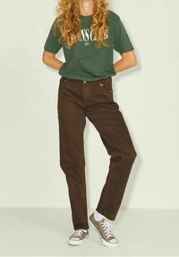 Jeans de algodón marrón con corte recto, combinados con una camiseta verde con texto blanco. Llevados con zapatillas gris y blancas.