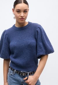 Navy blue knitted sweater with puff sleeves, crew neckline, and a textured finish, paired with high-waisted denim and a black belt.