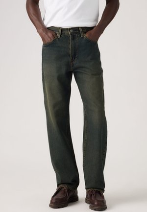 Bărbat purtând blugi verzi închiși cu picior drept, cămașă albă și pantofi maro din piele, cu mâinile în buzunare, stând pe un fundal simplu.