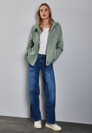 Veste légère vert clair avec capuche, chemise blanche et jeans bleus à jambes larges, complétés par des baskets blanches. Texture de tissu lisse.