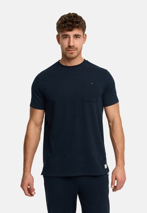 Jeune homme aux cheveux courts et bouclés portant une chemise bleu marine texturée à manches courtes avec une poche poitrine et un pantalon assorti.