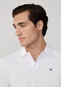 Junger Mann mit dunklen Haaren, der ein weißes Poloshirt mit einem kleinen gestickten Logo auf der linken Brust trägt und zur Seite schaut.