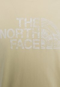 Krátký rukáv, světle béžové tričko s bílým logem s texturovanými horskými vzory; měkký bavlněný materiál s hladkým povrchem.