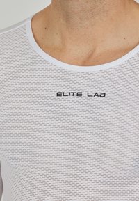 Hvid, tætsiddende atletisk skjorte lavet af mesh-stof, med rund halsudskæring og "ELITE LAB" logo i sort henover brystet.