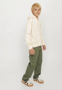 Krémová mikina s kapucí a přední kapsou, doplněná olivově zelenými cargo kalhotami s několika kapsami. Béžové tenisky s černými pruhy.