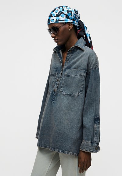 Άτομο με υπερμεγέθη τζιν πουκάμισο, ανοιχτόχρωμο παντελόνι, γεωμετρικό μπλε και λευκό μαντήλι στο κεφάλι και σκοτεινά γυαλιά ηλίου, κοιτάζοντας πλάγια με φόντο μονόχρωμο.