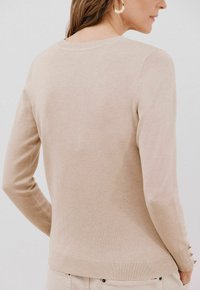 Beige Pullover mit langen Ärmeln aus weichem Strickstoff. Besitzt einen runden Halsausschnitt und eine gerippte Saum mit Knopfakzenten an den Ärmeln.