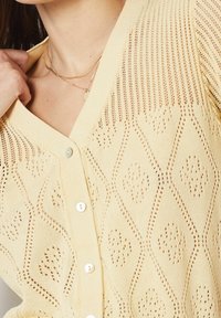 Cardigan de punto amarillo con patrones de diamantes y puntos, con un escote en V y botones blancos. La tela texturizada ofrece un diseño transpirable.