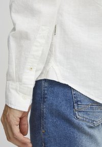 Camisa de lino blanca con puños abotonados, que presenta una textura suave y un ligero brillo, combinada con jeans de mezclilla azules.