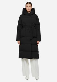 Cappotto imbottito nero con cappuccio oversized, vita regolabile con cintura e silhouette lunga. Presenta sezioni trapuntate e chiusura a bottoni.
