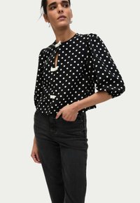 Blouse noire à pois avec des manches bouffantes, ouverture courte à l'avant et boutons décoratifs blancs, associée à un jean noir ajusté.