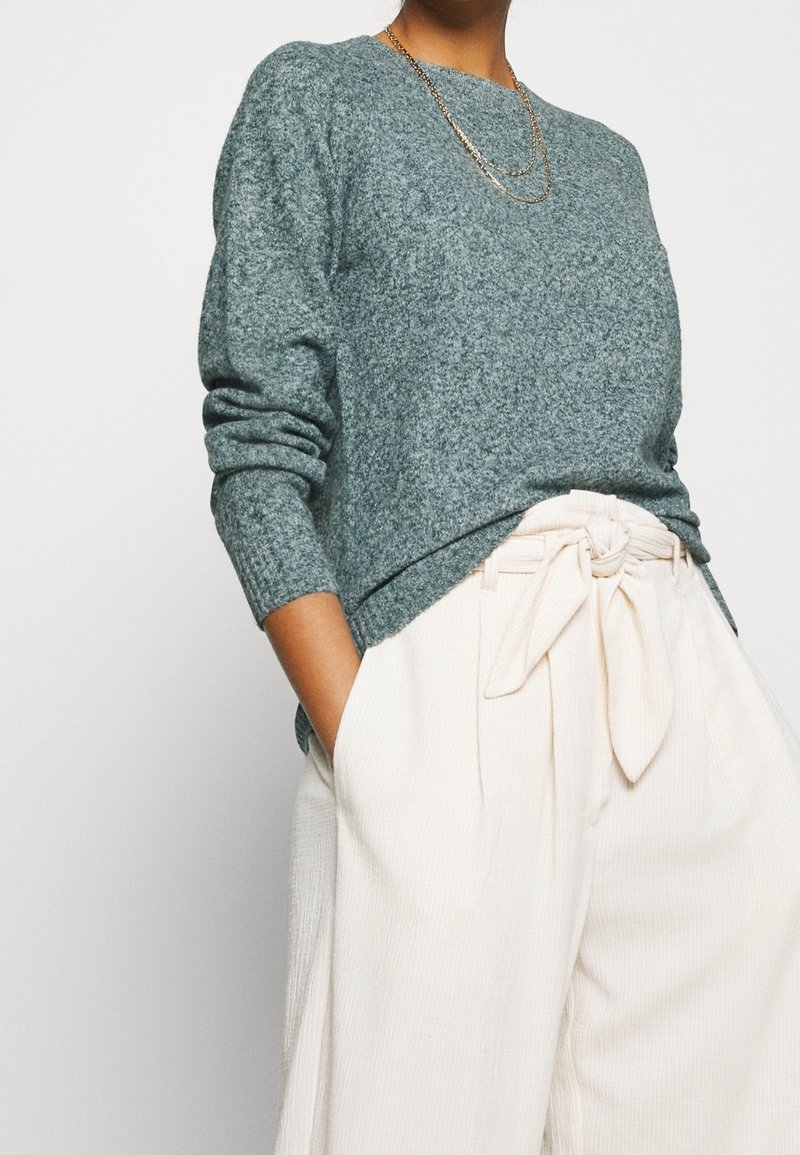 Personne portant un pull gris texturé à manches longues, un pantalon crème taille haute avec une ceinture nouée, et des colliers dorés superposés, les mains dans les poches.