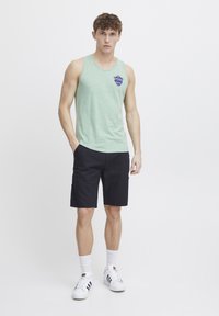 Top de tirantes verde heather con logotipo, combinado con pantalones cortos negros. El modelo lleva zapatillas blancas y calcetines hasta el tobillo. Diseño simple y casual.