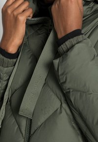 Zielona pikowana kurtka z zamkiem błyskawicznym z przodu, ściągaczami na mankietach oraz etykietą fabriczną z wytłoczonym napisem "Marc O'Polo" na uchwycie zamka.