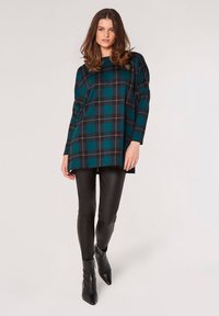 Oversized teal- und schwarze Karomuster-Tunika mit langen Ärmeln, kombiniert mit schwarzen engen Leggings und Ankle Boots, vor einem neutralen Hintergrund.