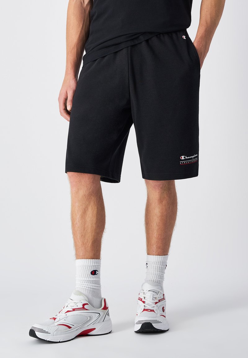 Zwarte sportshorts van een katoenmix, met een elastische tailleband. Heeft een klein "Champion" logo op de linker bovenbeen, gecombineerd met witte sokken en sneakers.