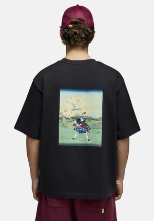 Personne portant un t-shirt noir avec un graphisme coloré d'un golfeur en tenue traditionnelle balançant un club sous un arbre, vue de dos.