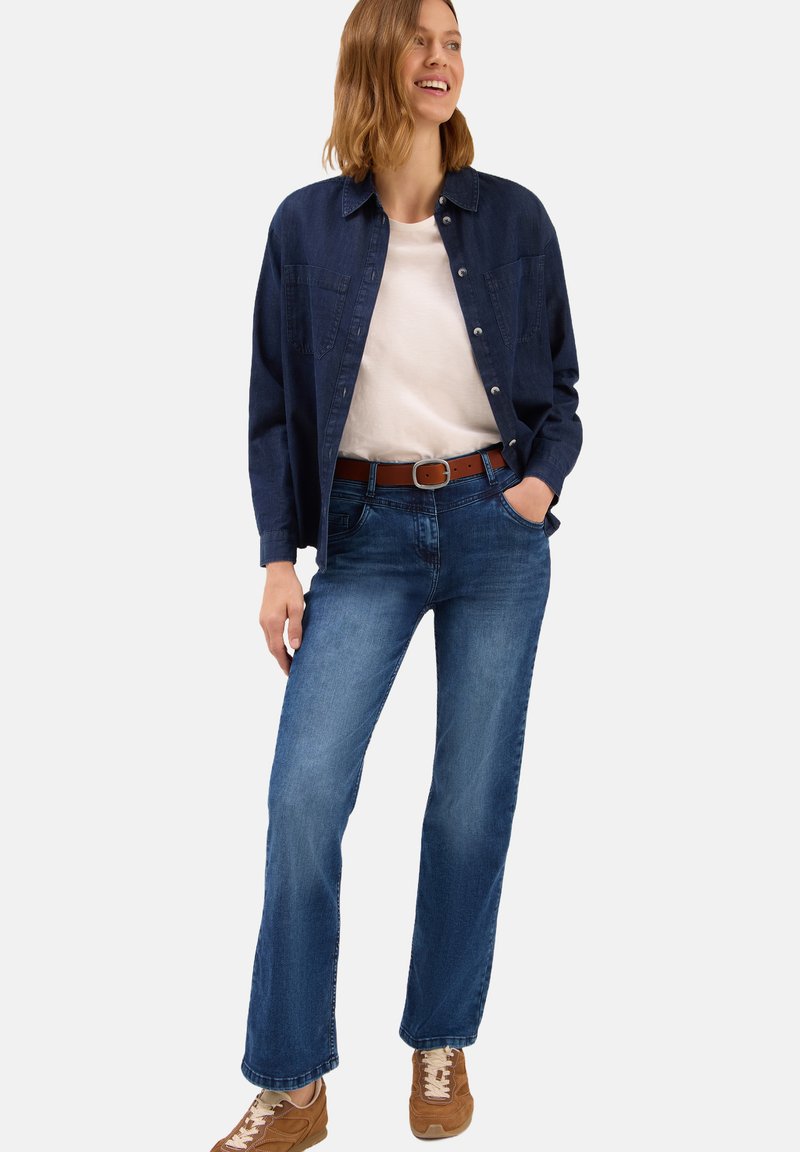 Blaue Jeans im geraden Schnitt, kombiniert mit einem weißen T-Shirt und einem dunkelblauen Hemd mit Knöpfen, ergänzt durch einen braunen Gürtel und beigen Sneakers.