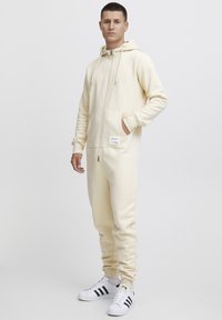 Tuta a zip color crema con cappuccio, tasche frontali e un'etichetta. Tessuto morbido, polsini aderenti e vestibilità rilassata complessiva. Indossata con sneakers bianche.