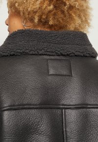 Giubbotto di pelle nera con colletto in shearling scuro, texture liscia con una toppa quadrata sul retro e dettagli di cucitura visibili.