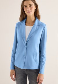Femme portant un blazer bleu clair boutonné sur un haut blanc et un jean gris foncé, debout devant un fond neutre uni.