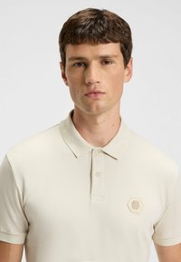 Beige polo shirt gemaakt van een gladde stof, met een gestructureerde kraag en een hexagonaal logo-opdruk op de linkerborst. Minimalistisch ontwerp.