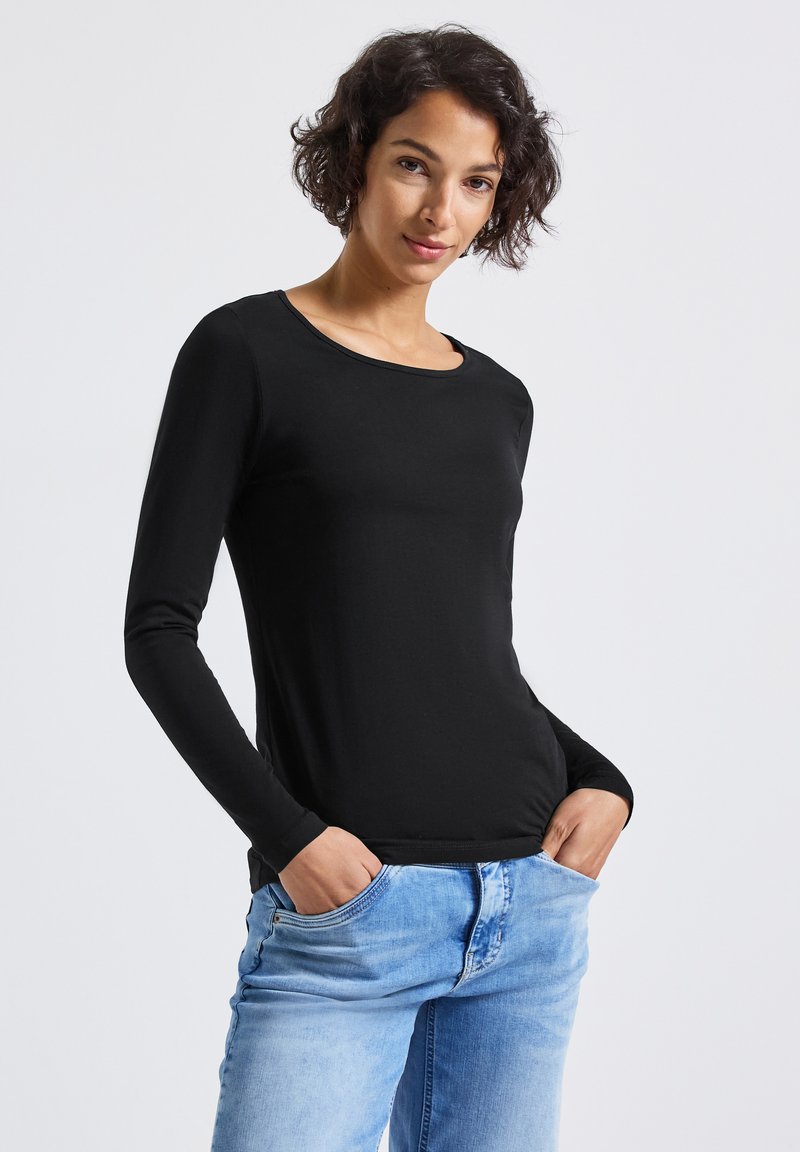 Donna con capelli corti e ricci che indossa una maglietta nera a maniche lunghe e jeans azzurri chiari, con le mani in tasca, in piedi davanti a uno sfondo semplice.
