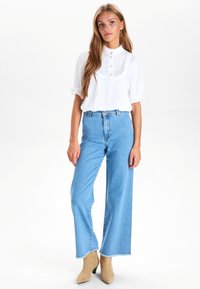 Vit blus med volangkrage och knappar, kombinerad med ljusblå högmidjade vida jeans och beige ankle boots.