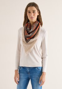 Jeune femme portant un haut beige à manches longues et un jean bleu, avec une écharpe marron et beige ornée de texte orange et d'accents imprimés léopard.