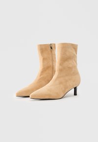 Beige semskede ankelstøvler med spidse tåer, sidelukning og slanke mellem-hæle på en ensfarvet hvit bakgrunn.