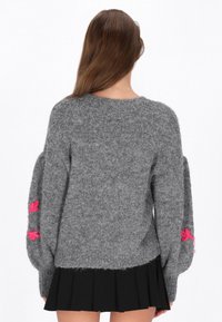 Grå stickad tröja med puffärmar, prydd med rosa blommig brodyr på varje ärm. Texturen är mjuk och fluffig, i kombination med en plisserad svart kjol.