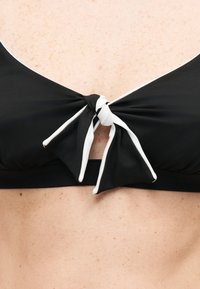 Parte superior de bikini negra con ribete blanco atada en un lazo en el centro sobre piel pálida y con pecas, mostrada desde el pecho hasta la parte superior del abdomen.