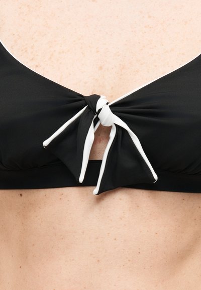 Μαύρο μπικίνι τοπ με λευκό τελείωμα δεμένο σε φιόγκο στο κέντρο σε ανοιχτόχρωμο, φακιδωτό δέρμα, εμφανίζεται από το στήθος μέχρι το άνω μέρος της κοιλιάς.