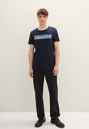 Camiseta de manga corta azul marino de algodón suave, con un logo impreso en blanco en el pecho y un cuello redondo. Combinada con pantalones y zapatos negros.
