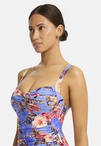 Een jonge vrouw met donker haar in een bun draagt een bloemenpatroon blauwe jurk, kijkt naar rechts met een neutrale uitdrukking.