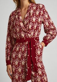 Haut enveloppant à motifs rouges avec des manches longues, présentant des formes de diamant en crème et jaune. Comprend une ceinture assortie et des poignets contrastés.