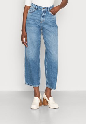 Person wearing light blue high-waist wide-leg cropped jeans and white heeled loafers, standing against a plain light background.