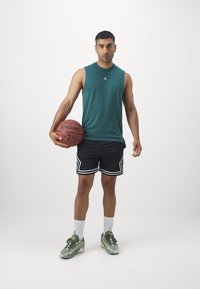 Zöld ujjatlan sportpóló, fekete rövidnadrág fehér csíkokkal, kosárlabdát tart, színes sportcipőt visel fehér zoknival.
