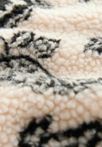 Strukturalna polarowa tkanina w jasnym beżu z czarnymi, kwiatowymi wzorami, charakteryzująca się miękką, puszystą powierzchnią oraz wypukłą, fluffową teksturą.
