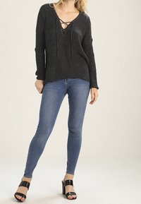 Maglione nero lavorato a maglia con scollatura a V profonda e dettagli con lacci, abbinato a jeans skinny blu e sandali neri con tacco a blocco.