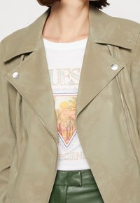 Veste en similicuir vert clair avec un col à revers, quincaillerie argentée et une fermeture éclair asymétrique, superposée sur un t-shirt graphique blanc.