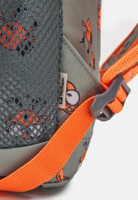 Gros plan sur un coin de sac gris et orange montrant une poche en maille, un motif de cartoon d'insecte orange, une sangle orange avec une boucle noire, et une étiquette de la marque Wolfskin.