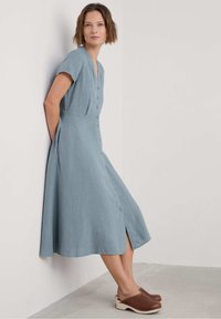 Abito in lino azzurro chiaro con scollatura a V, maniche corte, bottoni frontali, tasche laterali e orlo fino al ginocchio. Abbinato a zoccoli in legno marroni.
