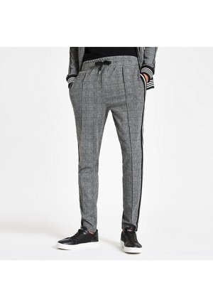 Harmaat ruudulliset jogger-housut mustilla sivuraitoja, yllään mustat tennarit ja vastaava ruudullinen takki, malli seisoo kädet taskuissa.