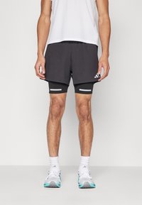 Schwarze Sportshorts mit grauen Akzenten, mehrlagiges Design, strukturierten Stoff, über engeren schwarzen Kompressionsshorts getragen, kombiniert mit weißen Sportschuhen.