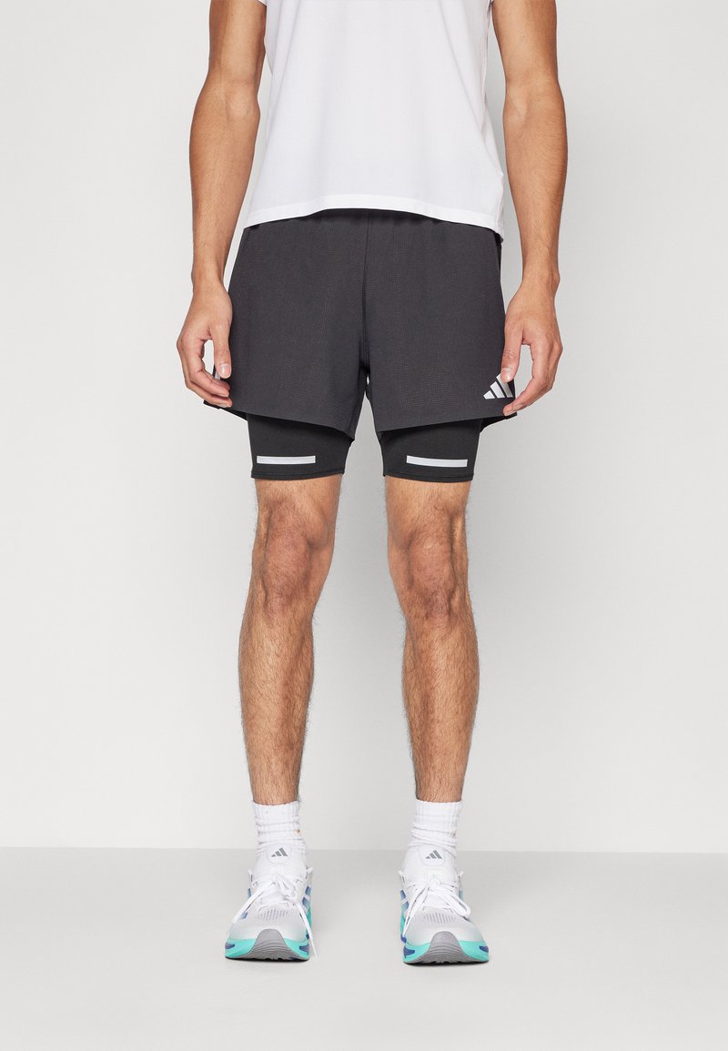 Schwarze Sportshorts mit grauen Akzenten, mehrlagiges Design, strukturierten Stoff, über engeren schwarzen Kompressionsshorts getragen, kombiniert mit weißen Sportschuhen.