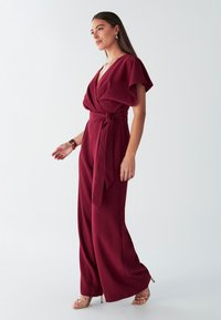 Mono burdeos con mangas cortas de vuelo, frente cruzado y cinturón en la cintura, con pantalones de pierna ancha y tacones dorados con tiras.