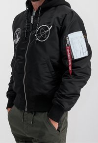 Musta bomber-takki, jossa on kirjailtuja avaruusteemaisia applikaatioita, resorit hihansuissa, vetoketjullinen hihatasku ja punainen "Remove Before Flight" -lappu.