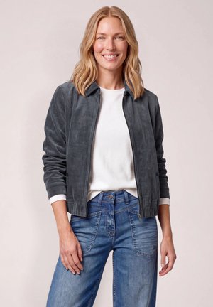 Mujer con cabello rubio hasta los hombros, que lleva una chaqueta gris con cremallera, camisa blanca y vaqueros azules, sonriendo con las manos relajadas a los lados.