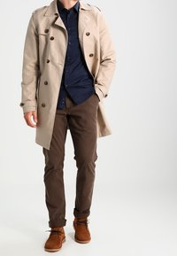 Beige trench coat with brown buttons, navy shirt underneath, brown trousers, and tan suede ankle boots. The coat has a classic cut and collar.